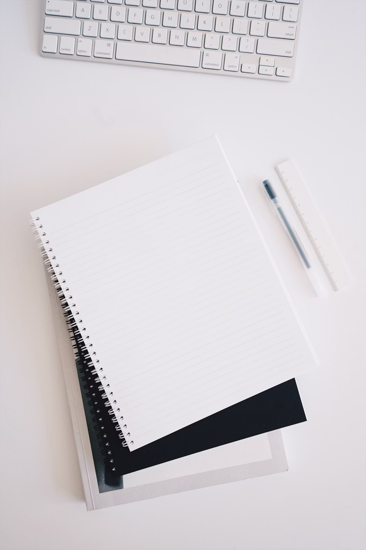 Minimal workspace with a spiral notebook, keyboard, pen, and ruler arranged on a clean white desk.